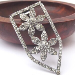 May include: A silver-toned brooch with a floral design. Two flower shapes are set with clear rhinestones, within a rectangular frame also set with rhinestones. The brooch is set against a wooden background.
