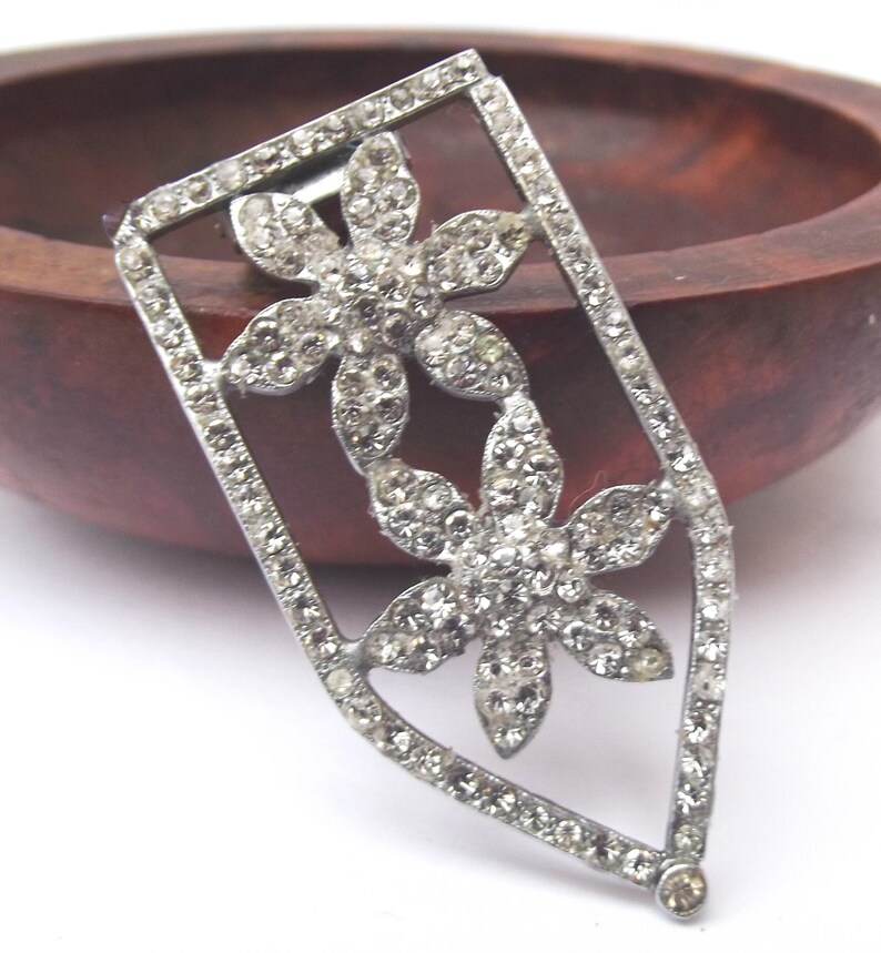 May include: A silver-toned brooch with a floral design. Two flower shapes are set with clear rhinestones, within a rectangular frame also set with rhinestones. The brooch is set against a wooden background.