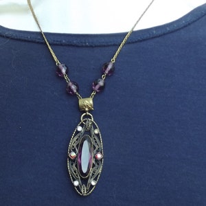 May include: A purple glass pendant necklace with a filigree design and a large oval-shaped purple gemstone. The necklace is made of a gold-toned chain with small purple glass beads.
