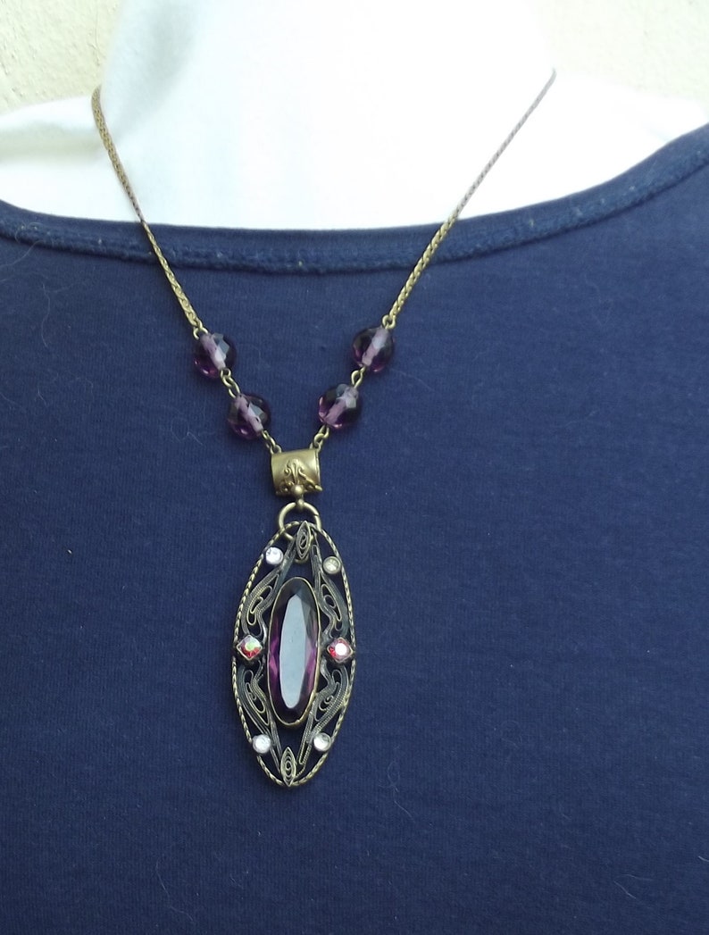 May include: A purple glass pendant necklace with a filigree design and a large oval-shaped purple gemstone. The necklace is made of a gold-toned chain with small purple glass beads.