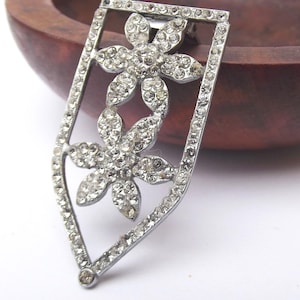 May include: A silver-toned brooch with a shield-like frame, encasing two flower designs. Each flower is adorned with numerous clear, sparkling rhinestones. The brooch has a pin closure on the back, suitable for attaching to clothing or accessories.