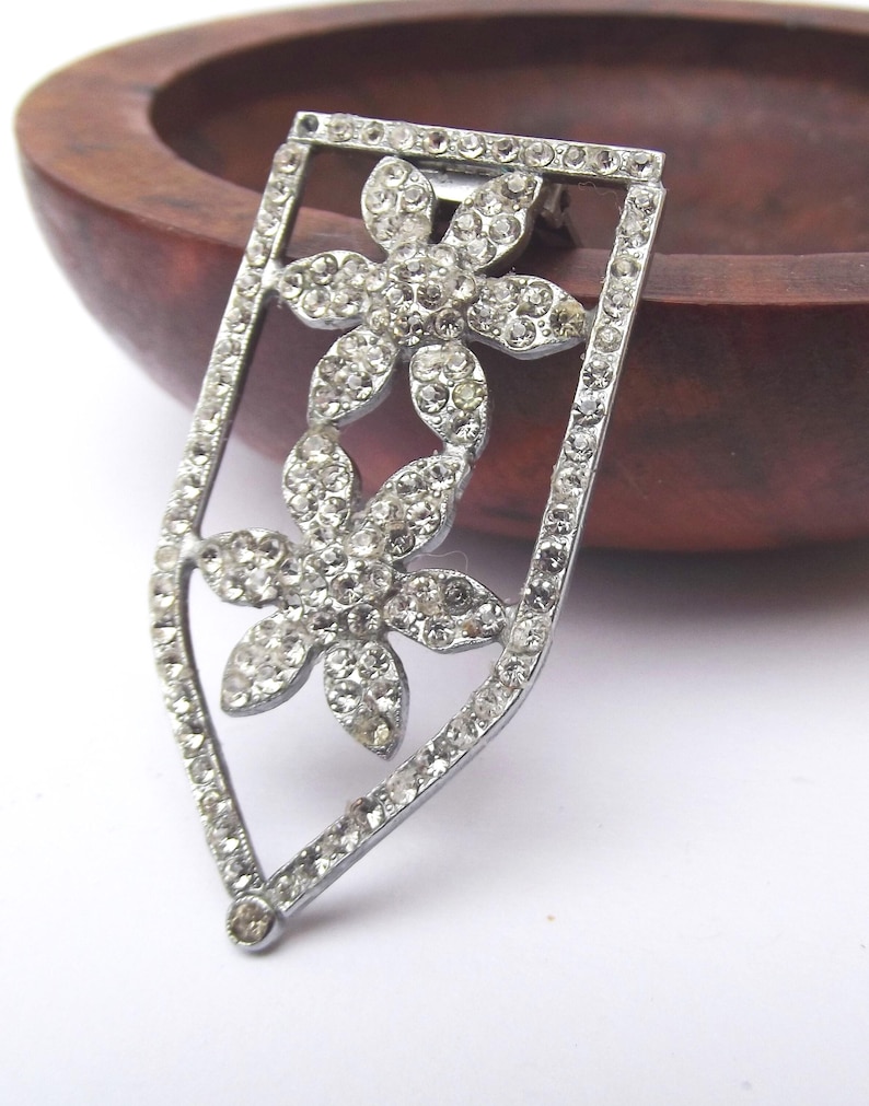 May include: A silver-toned brooch with a shield-like frame, encasing two flower designs. Each flower is adorned with numerous clear, sparkling rhinestones. The brooch has a pin closure on the back, suitable for attaching to clothing or accessories.