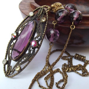May include: A vintage-style pendant necklace with a large, oval-shaped amethyst stone set in an ornate, filigree frame. The necklace features a chain made of small, linked brass pieces and is adorned with additional amethyst beads.