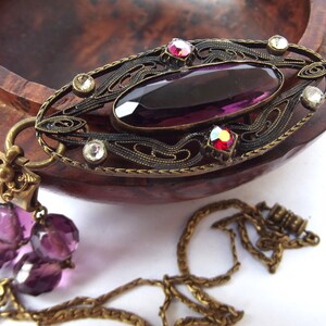 May include: A vintage pendant necklace with a large, oval-shaped, purple glass stone set in an ornate, filigree frame. The frame is made of brass and has a detailed design with small, clear rhinestones. The necklace has a chain with a purple glass bead tassel.
