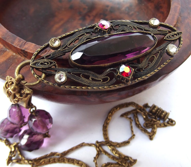 May include: A vintage pendant necklace with a large, oval-shaped, purple glass stone set in an ornate, filigree frame. The frame is made of brass and has a detailed design with small, clear rhinestones. The necklace has a chain with a purple glass bead tassel.