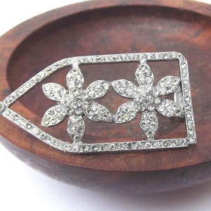 May include: A silver-toned vintage brooch featuring two flower designs encrusted with clear rhinestones. The brooch is rectangular with a pointed end and is set against a wooden background.