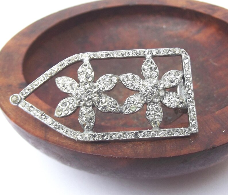 May include: A silver-toned vintage brooch featuring two flower designs encrusted with clear rhinestones. The brooch is rectangular with a pointed end and is set against a wooden background.