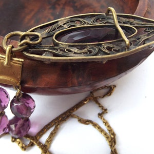May include: A gold chain necklace with a pendant featuring a filigree design and a large purple glass stone. The pendant is attached to the chain with a gold clasp.