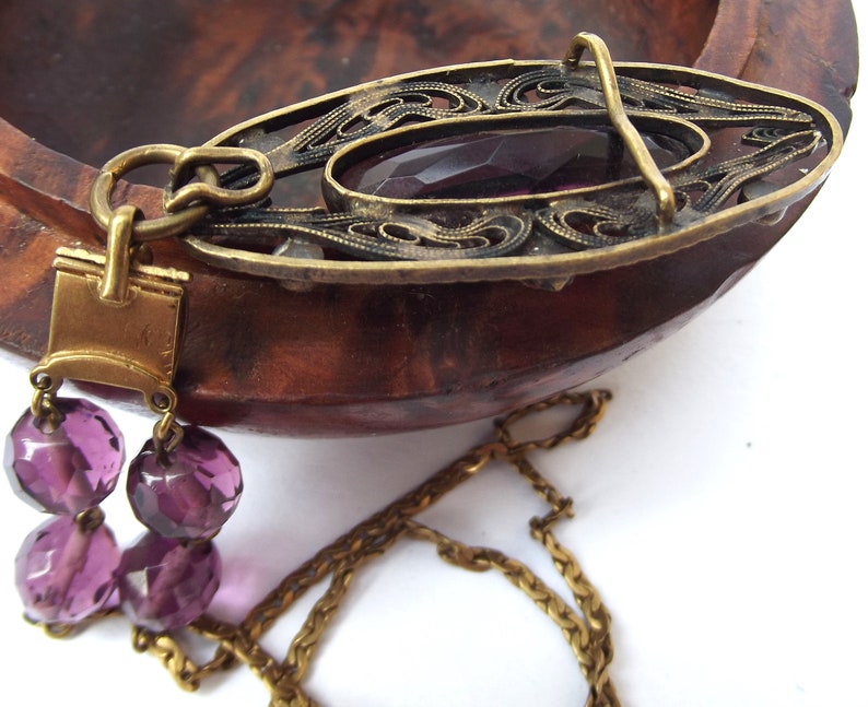 May include: A gold chain necklace with a pendant featuring a filigree design and a large purple glass stone. The pendant is attached to the chain with a gold clasp.