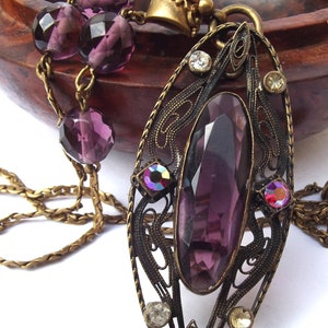 May include: A vintage-style pendant necklace with a large, faceted purple gemstone set in an ornate, filigree frame. The pendant is attached to a chain with a series of smaller purple beads.