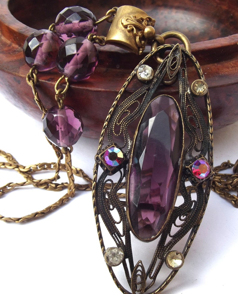 May include: A vintage-style pendant necklace with a large, faceted purple gemstone set in an ornate, filigree frame. The pendant is attached to a chain with a series of smaller purple beads.