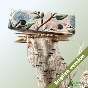 May include: A beige fabric needle case with a floral pattern featuring blue flowers, green leaves, and pink buds, secured by two black snaps and embroidered with the letters AS. It holds multiple metallic knitting needles and a partially knitted sock.