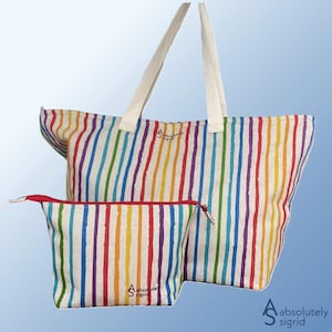 Op de afbeelding: Een grote draagtas en een kleinere make-up tas, beide met een regenboog gestreept patroon. De tassen hebben witte handvatten en een rode rits op de kleinere tas. De tekst "absolutely sigrid" is op beide tassen gedrukt.