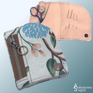 May include: Two fabric cases for sewing supplies. One is a peach-colored needle case with needles and a pair of rainbow scissors. The other is a zippered pouch with a floral pattern in blue, green, and pink. The pouch has a brown zipper.