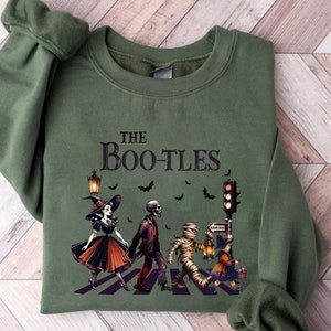 May include: Green sweatshirt with the text "The Bootles" and a Halloween-themed graphic. The graphic features a witch, a skeleton, a mummy, and a trick-or-treater crossing a crosswalk. Bats and a traffic light are also included.