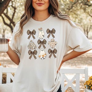 A woman wearing a white t-shirt with a graphic of ghostly figures and bows stands in a park surrounded by pumpkins and hay bales.