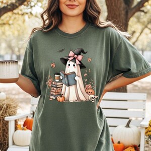 A woman wearing a green t-shirt with a graphic of a ghost reading a book, surrounded by Halloween decorations such as pumpkins, gourds, and a mug.