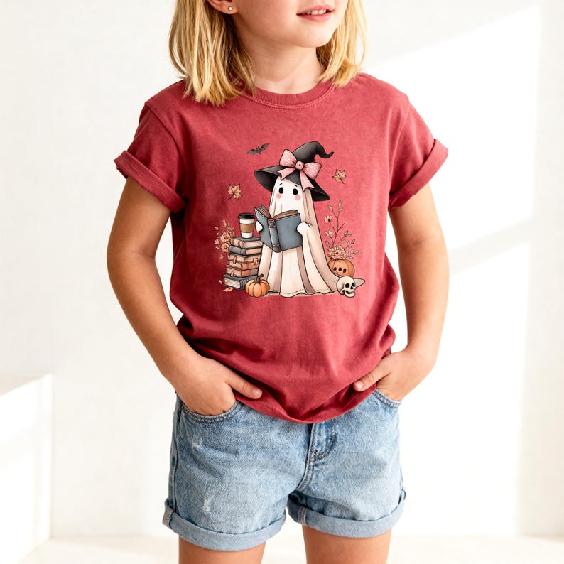 A young girl wearing a red t-shirt with a cartoon ghost character reading a book, surrounded by Halloween-themed decorations.