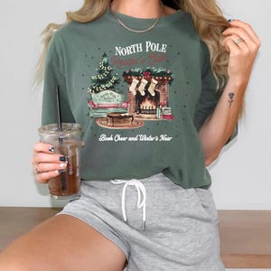 A woman wearing a green t-shirt with a Christmas-themed graphic and holding a drink, smiling at the camera.