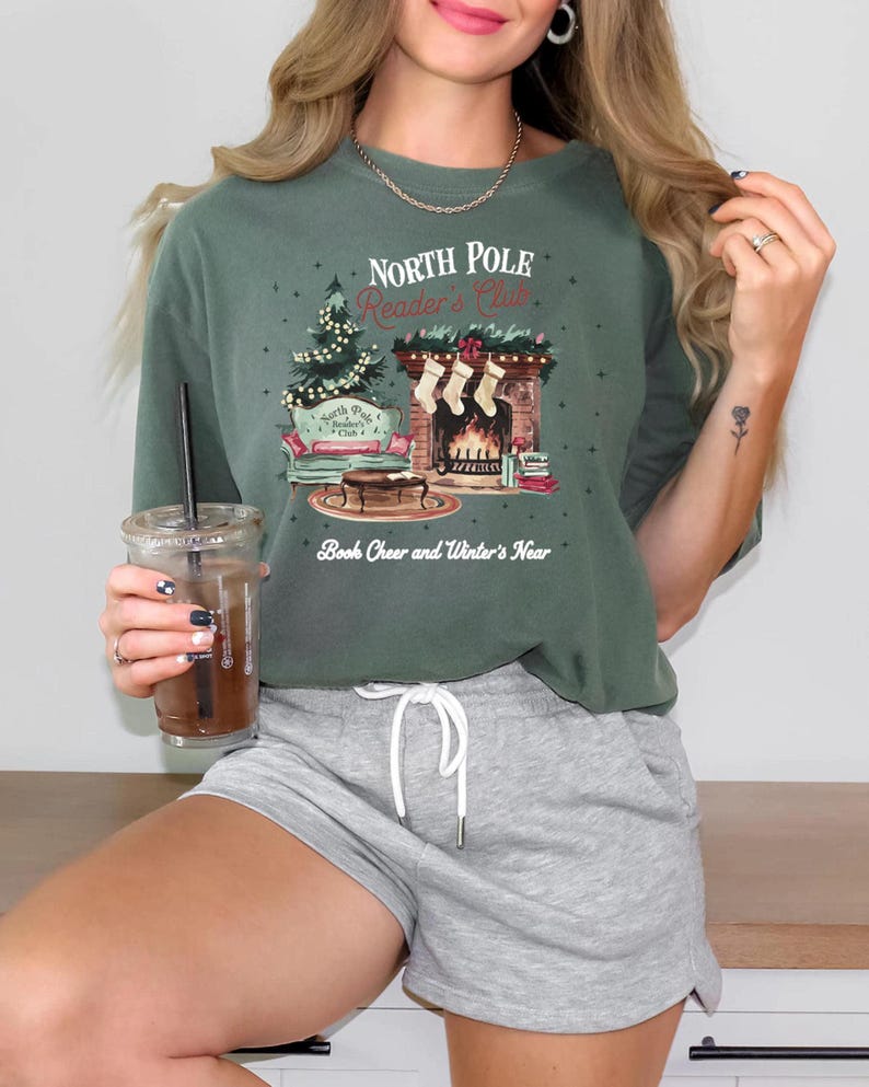 A woman wearing a green t-shirt with a Christmas-themed graphic and holding a drink, smiling at the camera.