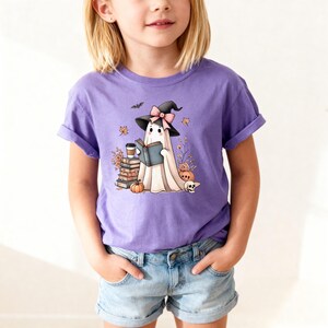 A young girl wearing a purple t-shirt with a graphic of a ghost reading a book and surrounded by Halloween-themed decorations.