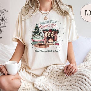 A woman wearing a white t-shirt with a graphic design of a cozy winter scene, including a fireplace, a Christmas tree, and a book, is sitting on a bed with a white blanket. She is holding a white mug in her hand.