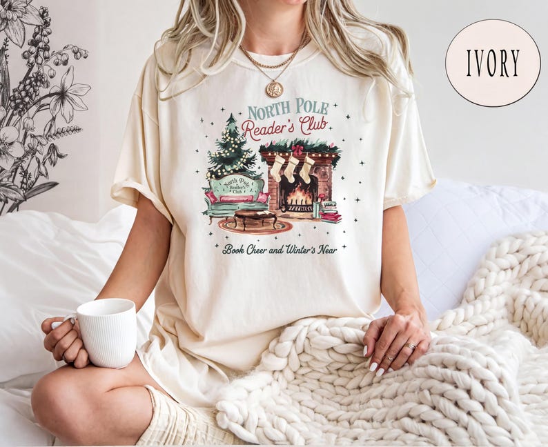A woman wearing a white t-shirt with a graphic design of a cozy winter scene, including a fireplace, a Christmas tree, and a book, is sitting on a bed with a white blanket. She is holding a white mug in her hand.