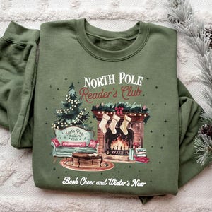 A green sweatshirt with a Christmas-themed design, including a fireplace, Christmas tree, and stockings, along with the text "North Pole Reader's Club" and "Book Cheer and Winter's Near".
