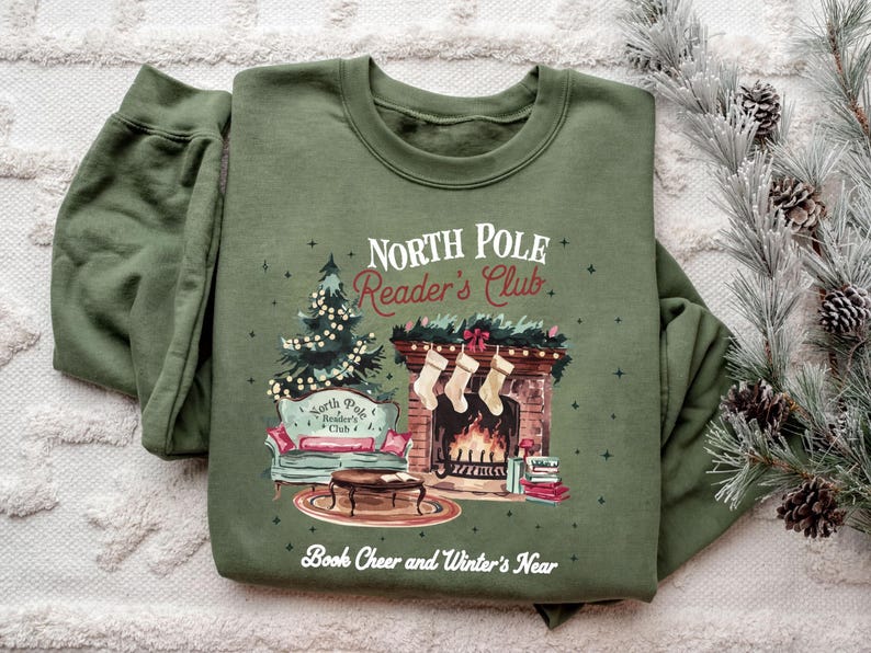 A green sweatshirt with a Christmas-themed design, including a fireplace, Christmas tree, and stockings, along with the text "North Pole Reader's Club" and "Book Cheer and Winter's Near".