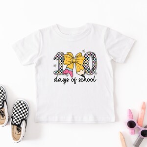 A white t-shirt with a graphic design, a pair of black and white checkered shoes, and some children's school supplies like pencils and sunglasses are arranged on a white background.