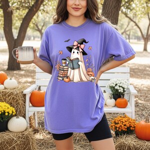 A woman wearing a purple t-shirt with a graphic of a ghost reading a book, holding a mug, and surrounded by pumpkins, flowers, and a bench in a park-like setting.