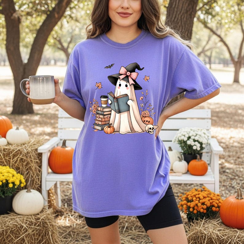A woman wearing a purple t-shirt with a graphic of a ghost reading a book, holding a mug, and surrounded by pumpkins, flowers, and a bench in a park-like setting.