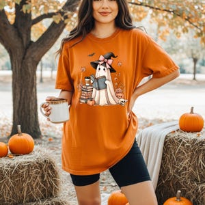 A woman wearing an orange t-shirt with a graphic of a ghost holding a book and a mug, standing in front of hay bales and pumpkins.
