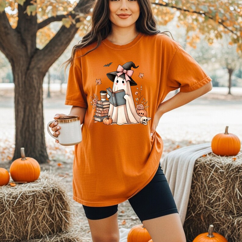 A woman wearing an orange t-shirt with a graphic of a ghost holding a book and a mug, standing in front of hay bales and pumpkins.