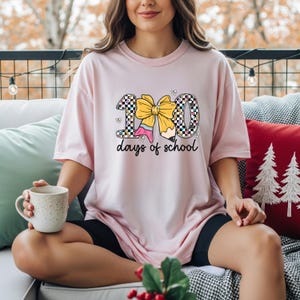 A woman sitting on a couch, wearing a pink t-shirt with a graphic design of a yellow bow and the text "100 days of school". She is holding a coffee mug in her hand.