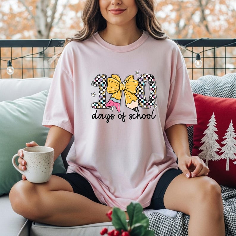 A woman sitting on a couch, wearing a pink t-shirt with a graphic design of a yellow bow and the text "100 days of school". She is holding a coffee mug in her hand.