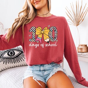 A woman wearing a red sweatshirt with a graphic design of a yellow bow and the text "100 days of school" is sitting on a bed.