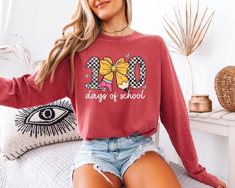 A woman wearing a red sweatshirt with a graphic design of a yellow bow and the text "100 days of school" is sitting on a bed.