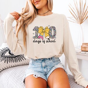 A woman wearing a white long-sleeved shirt with a graphic design of a yellow bow and the text "100 days of school" is sitting on a bed.