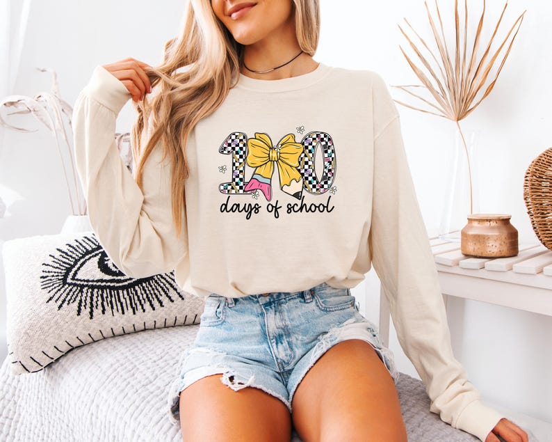 A woman wearing a white long-sleeved shirt with a graphic design of a yellow bow and the text "100 days of school" is sitting on a bed.
