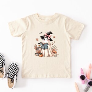 A beige t-shirt with a graphic of a ghost wearing a witch's hat and holding a book, surrounded by Halloween-themed elements such as pumpkins, bats, and a skull.