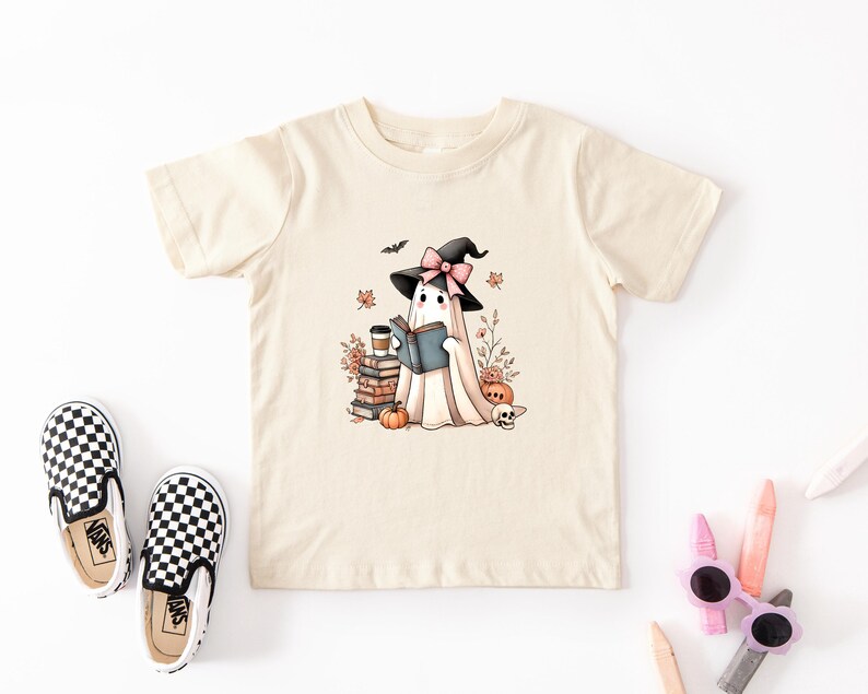 A beige t-shirt with a graphic of a ghost wearing a witch's hat and holding a book, surrounded by Halloween-themed elements such as pumpkins, bats, and a skull.