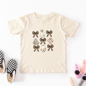 a beige t-shirt with a graphic design of ghostly figures with bows, a pair of black and white checkered shoes, and some pink and purple markers.