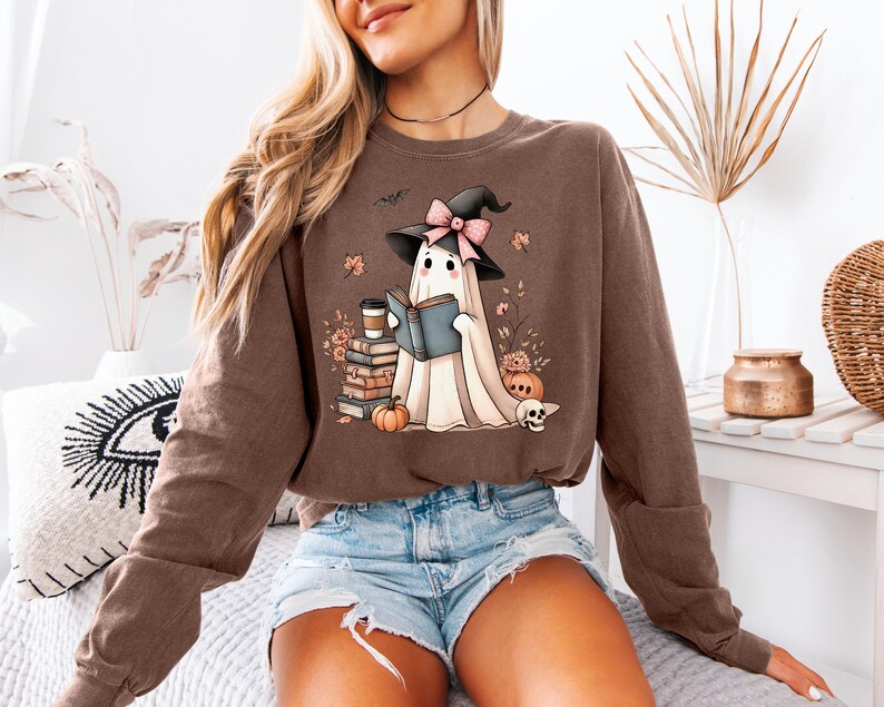 A woman wearing a brown sweatshirt with a graphic of a ghost reading a book, surrounded by Halloween-themed elements such as pumpkins, bats, and a skull.