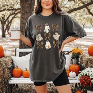 A woman wearing a black t-shirt with a graphic of ghostly figures and leopard-print bows, holding a mug, and standing in front of a Halloween-themed arrangement with pumpkins, flowers, and hay bales.