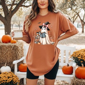 A woman wearing a brown t-shirt with a graphic of a ghost holding a book and a mug, standing in front of a white bench with pumpkins and hay bales.