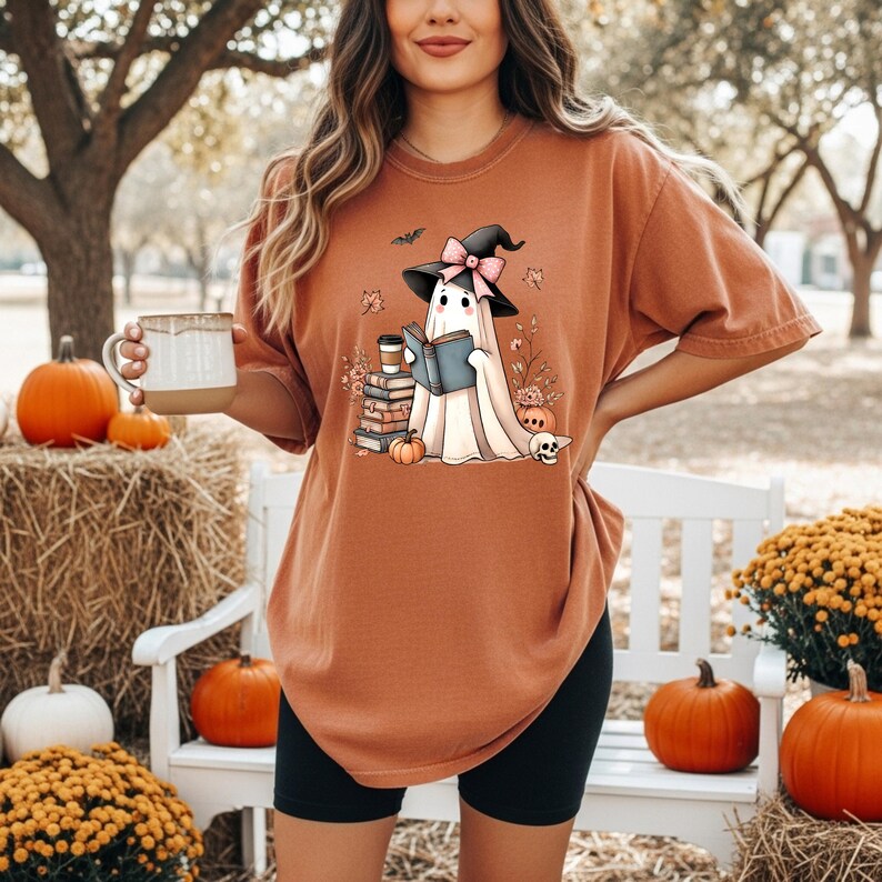 A woman wearing a brown t-shirt with a graphic of a ghost holding a book and a mug, standing in front of a white bench with pumpkins and hay bales.