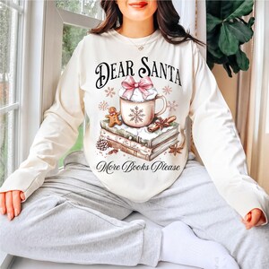 A woman wearing a white sweater with a graphic design of a mug, books, and a snowflake, sitting in a window with a potted plant in the background.