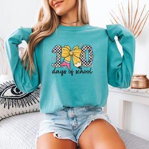 A woman wearing a teal long-sleeved shirt with a graphic design and text, sitting on a bed with a decorative pillow.