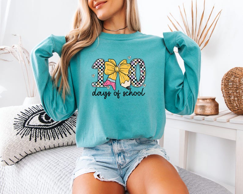 A woman wearing a teal long-sleeved shirt with a graphic design and text, sitting on a bed with a decorative pillow.